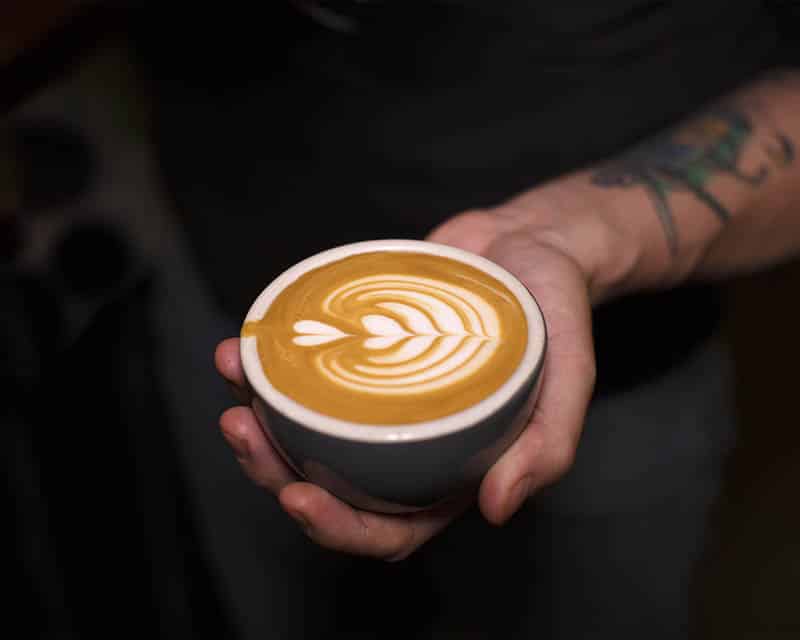 tattoed arm and hand cradling cup of coffee with latte art of hearts