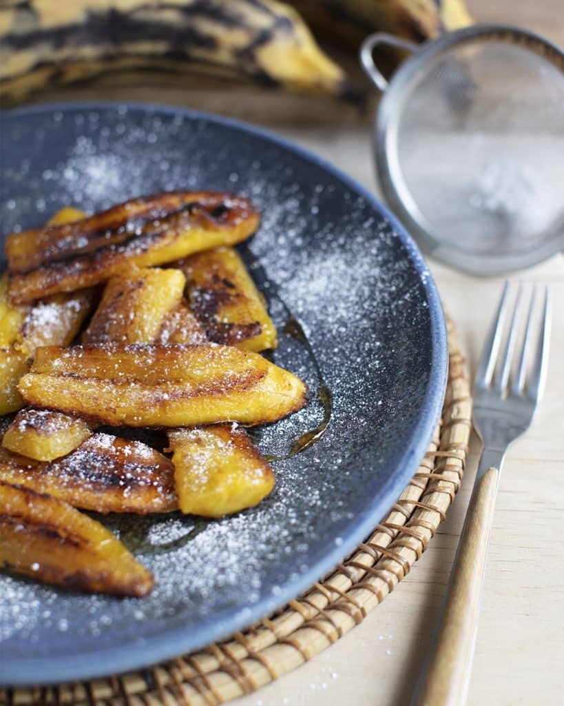 beautiful food photography of fried plantain sprinkled with icing sugar on a blue plate with sieve and fork in background